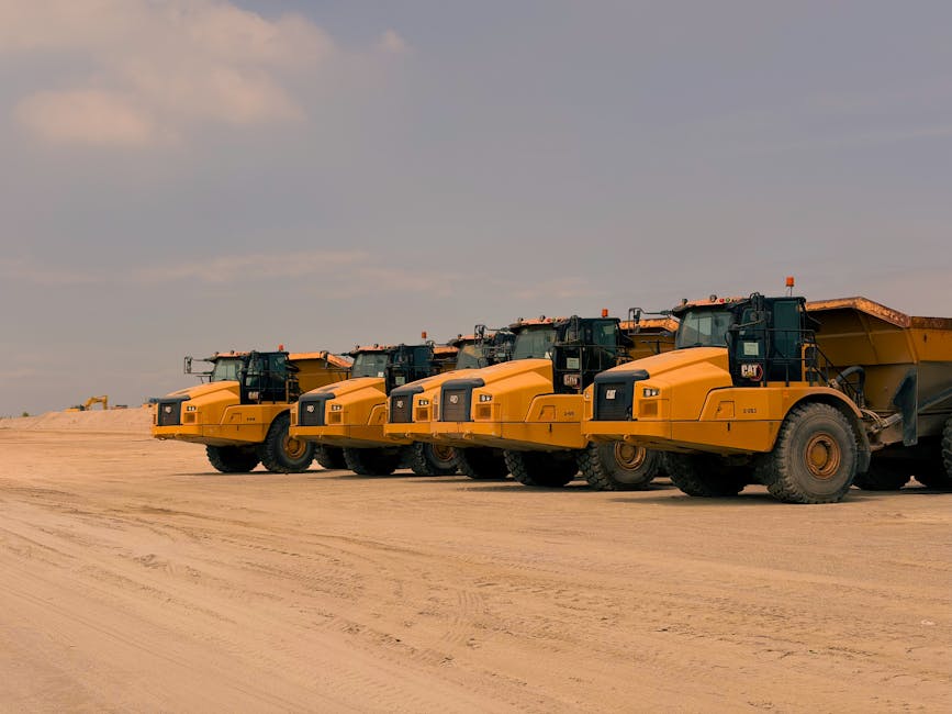 Row of heavy-duty dump trucks parked on a sandy construction site under a clear sky.